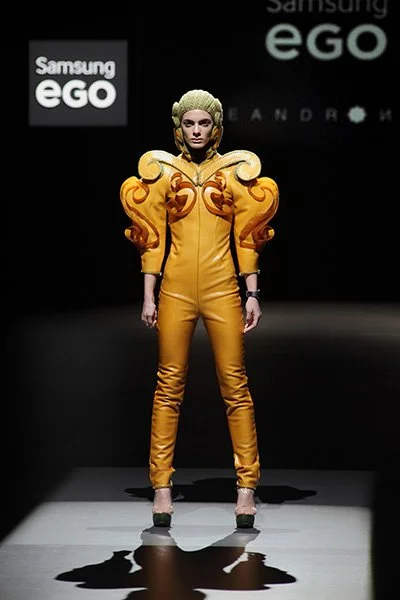 Model wearing a mustard fitted suit with exaggerated curled shoulders from 083