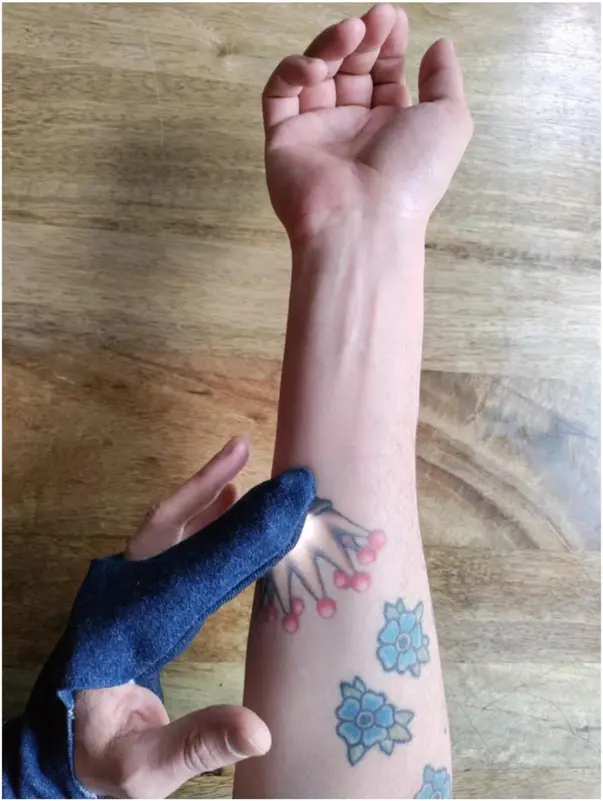 The illuminated fingertip of the denim glove sensing colour on a tattooed forearm.