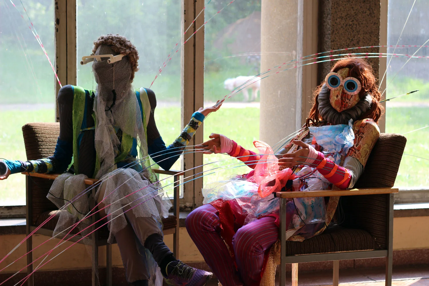 Two performers seated in layered costumes connected to long flexible rods