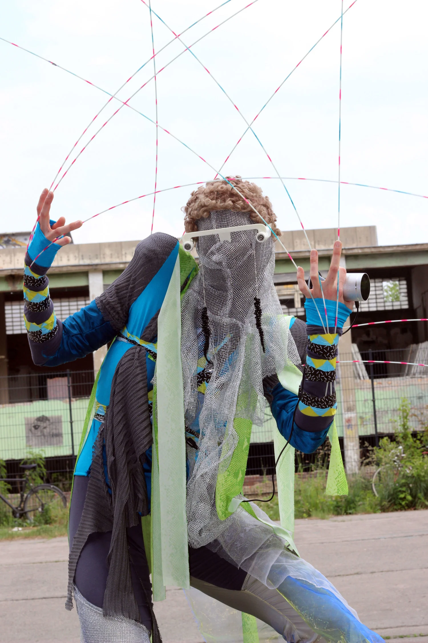 A masked performer in a blue, green and grey wearable structure surrounded by long flexible rods