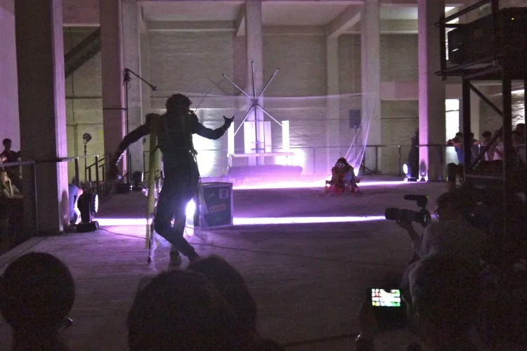 A silhouetted performer moving with extended rods before an audience in a dark industrial hall